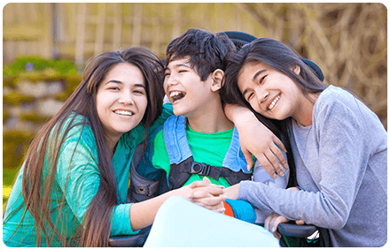 three siblings are in a group hug, smiling and laughing together. the boy in the middle is using a wheelchair.