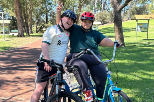 Two Rocky Bay supported accommodation customers are on bicycles. They are standing next to each other and smiling. They are both wearing helmets and the customer on the right has his arm around the customer on the left.