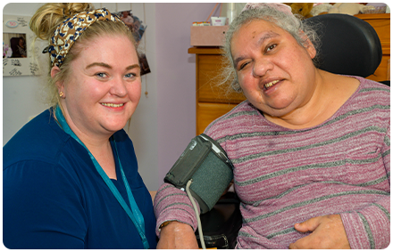 a rocky bay registered nurse sits and smiles with a rocky bay client who lives with a disability. the nurse is checking the client's blood pressure at home.
