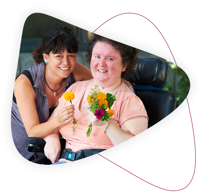 a disability support worker and a person living with disability and is using a wheelchair smile together. they are both holding flowers.