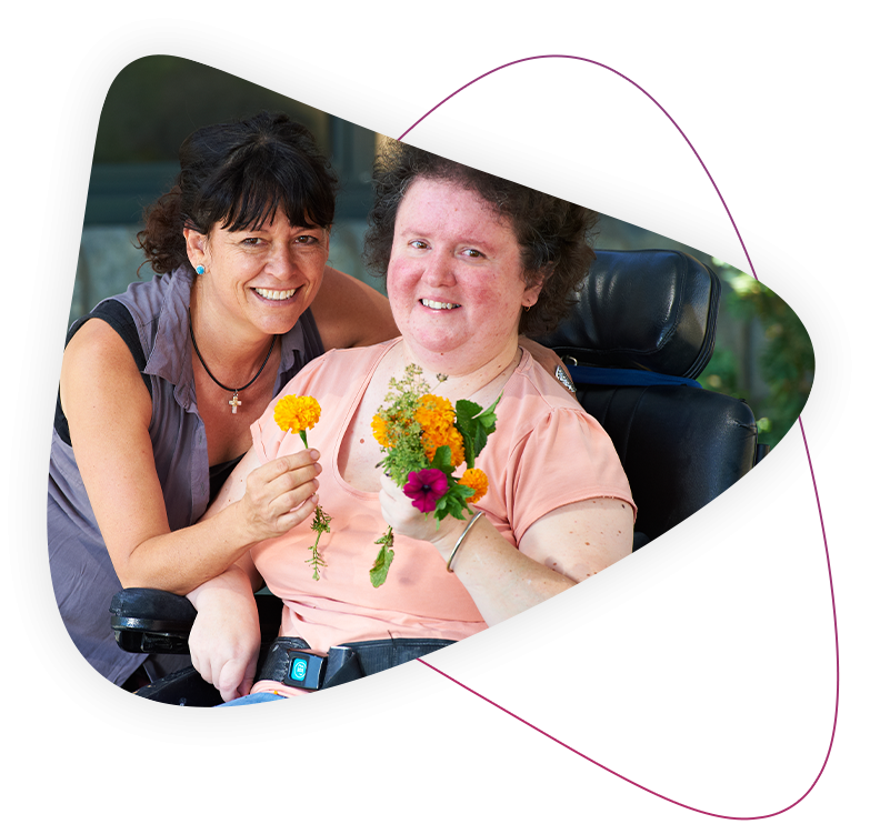 About Rocky Bay - Mosman Park a disability support worker and a person living with disability and is using a wheelchair smile together. they are both holding flowers.