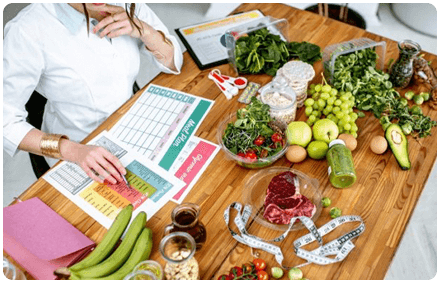 dietitian wears a white shirt and looks at colourful charts on documents with fresh food and a measuring tape on the desk surrounding the dietitian.