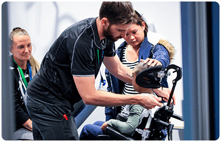 a rocky bay occupational therapist assists the neck of a child who lives with a disability. the child sits in a wheelchair. his family member places a hand to the top of his head.