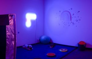 Photo showing the sensory room at the Rocky Bay Joondalup hub. here is a blue neon light, lots of soft toys, a tent and reflective shapes on the walls.