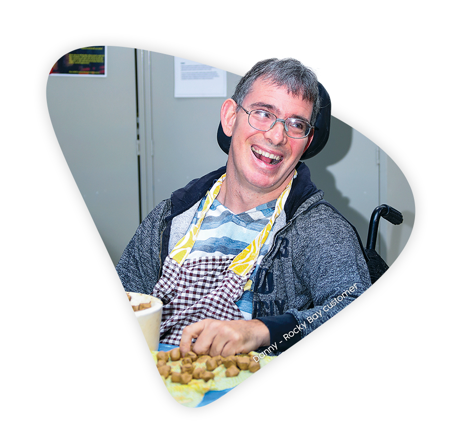 danny, a rocky bay customer, cooks and smiles while he sits in a rocky bay wheelchair. he wears glasses and an apron on top of his casual clothes.