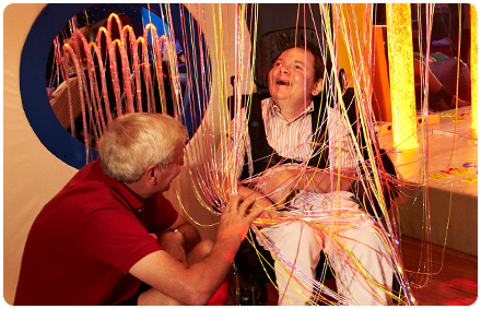 a man who is using a wheelchair sits under fibre optic lights with a rocky bay support worker in a sensory room.