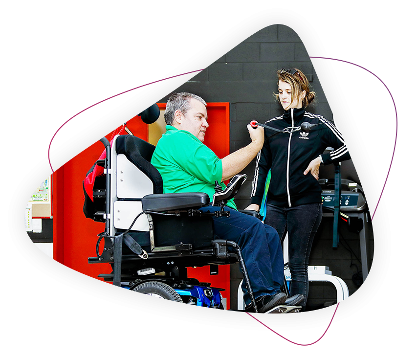 a man who lives with a disability is sitting in his wheelchair and working out at the gym. there is a rocky bay physiotherapist helping him.