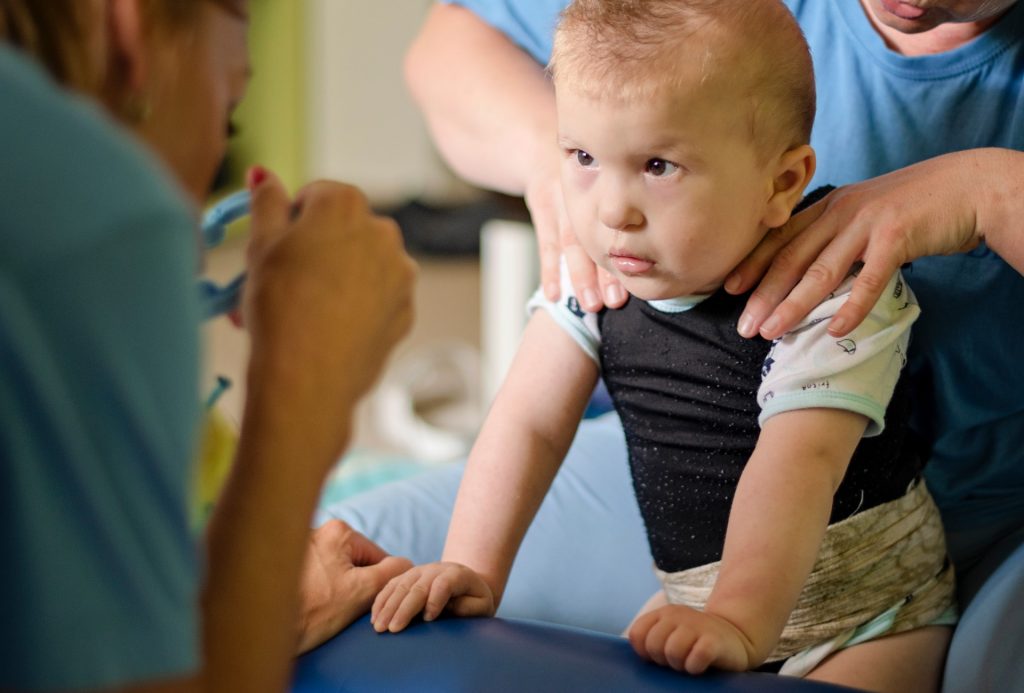 infant participates in the early start intervention program. he sits on a medical bed while a rocky bay medical professional treats his back. another rocky bay medical professional engages with the child.