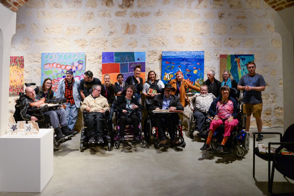 A group photo taken at the WA Shipwrecks Museum of Rocky Bay's artists and the support workers from the Studio, with colourful artworks in the background.