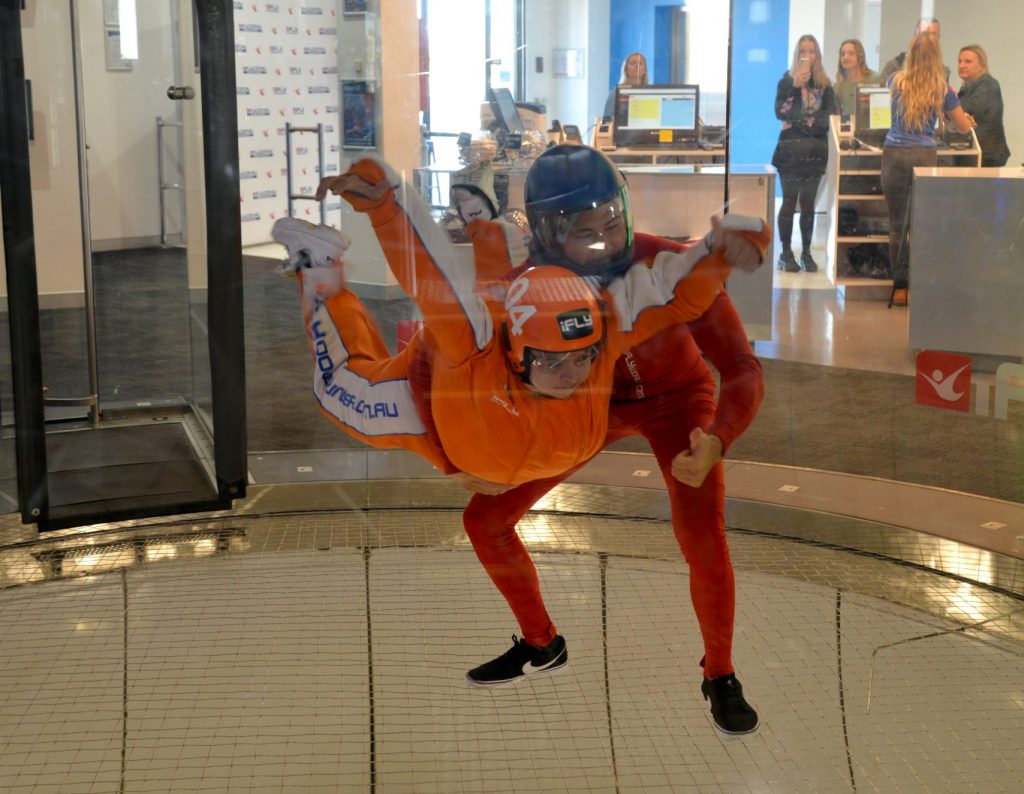 Fenton experiencing indoor skydiving with a professional instructor, wearing an orange skydive outfit