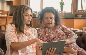 two women are sitting together on a couch and are looking at a tablet device that one of the women is holding. She is pointing at the ipad.