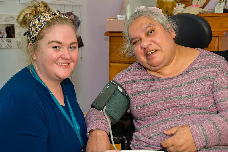 Rocky Bay Nurse, Louise, in a blue shirt and smiling is sitting next to a smiling Rocky Bay customer whilst taking her blood pressure