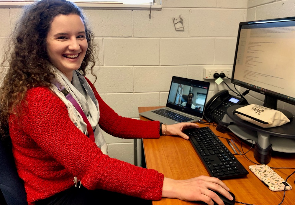 Cloe sits at her desk conducting a telepractice session. She wears a bright red jumper and is smiling at the camera