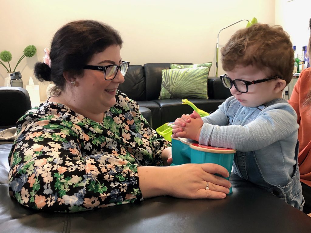 Young customer Henry and his mum Leanne. Henry is wearing a blue tshirt and denim overalls and is playing with a switch toy. Leanne is smiling and wearing a floral blouse.