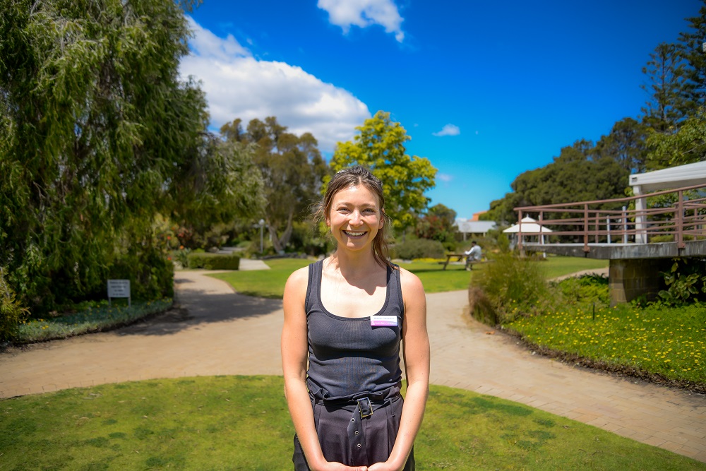 Mariah wears a black t-shirt and is standing in the gardens of Rocky Bay in Mosman Park