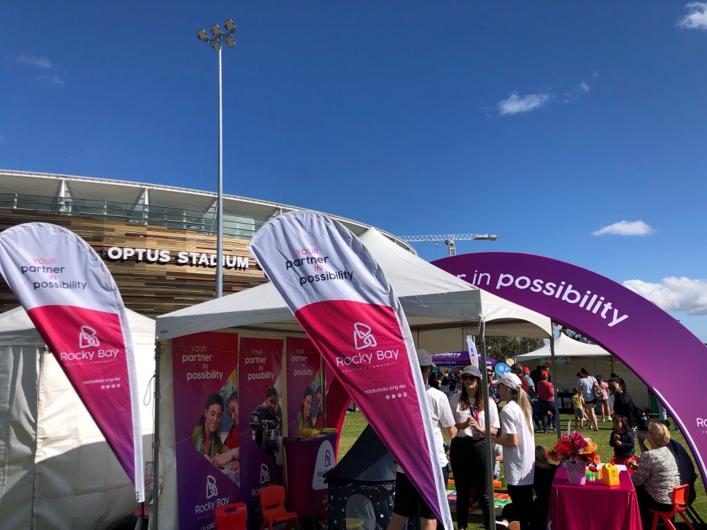Rocky Bay's marquee set up during Telethon Family Festival. The marquee is filled with Rocky Bay banners and flags Optus Stadium is in the background.