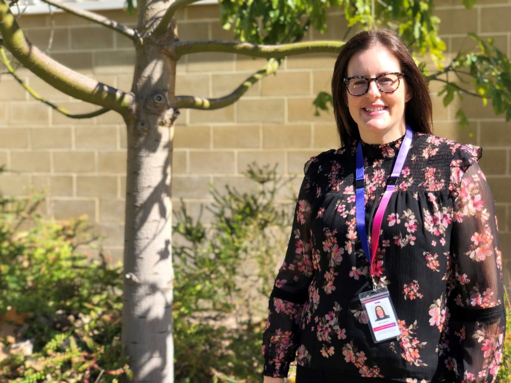 Ellie is standing in the gardens of Rocky Bay's Mosman Park hub. She is wearing a black and pink floral blouse.