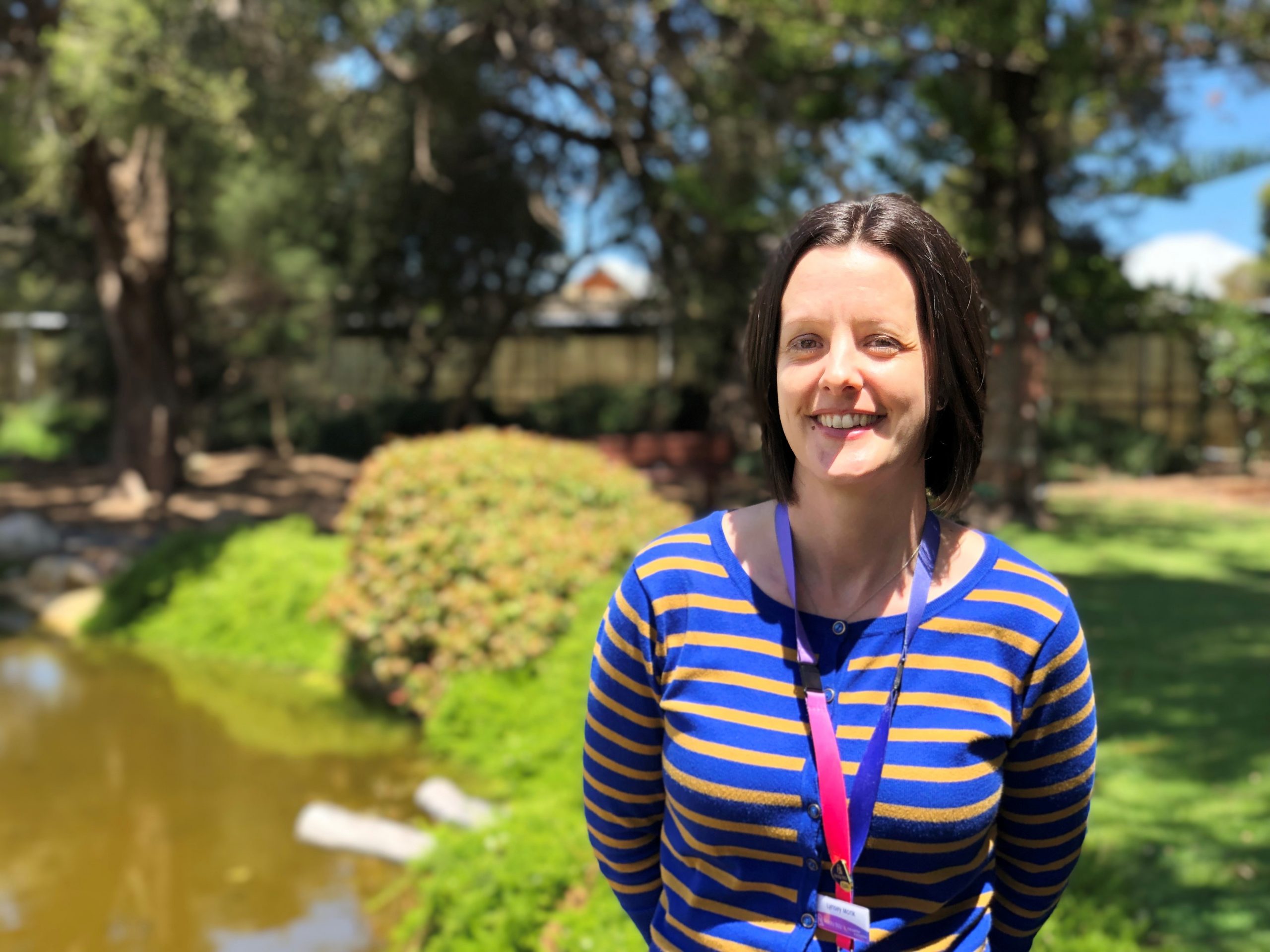 Lynsey is standing in the gardens of Mosman Park. She is smiling at the camera and wearing a blue striped top