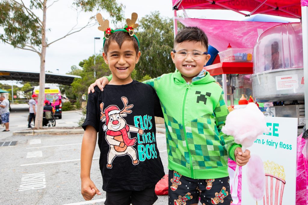 Two young boys at the Family Fun Day are smiling. One is wearing a black tshirt and the other a green tshirt with his arm around the other. The boy in green is holding fairy floss