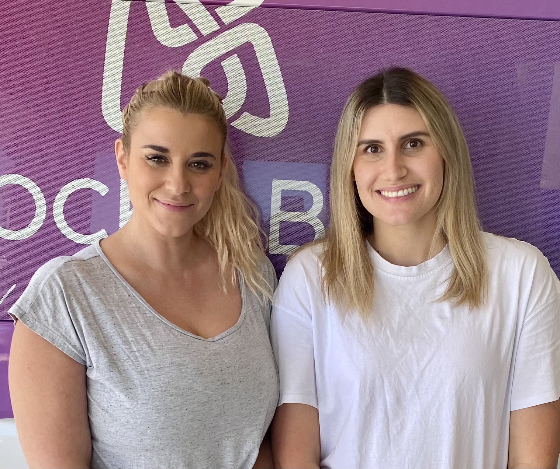 Sam is wearing a grey t-shirt and is standing next to and Ash, who is wearing a white t-shirt. There is pink Rocky Bay signage behind them.