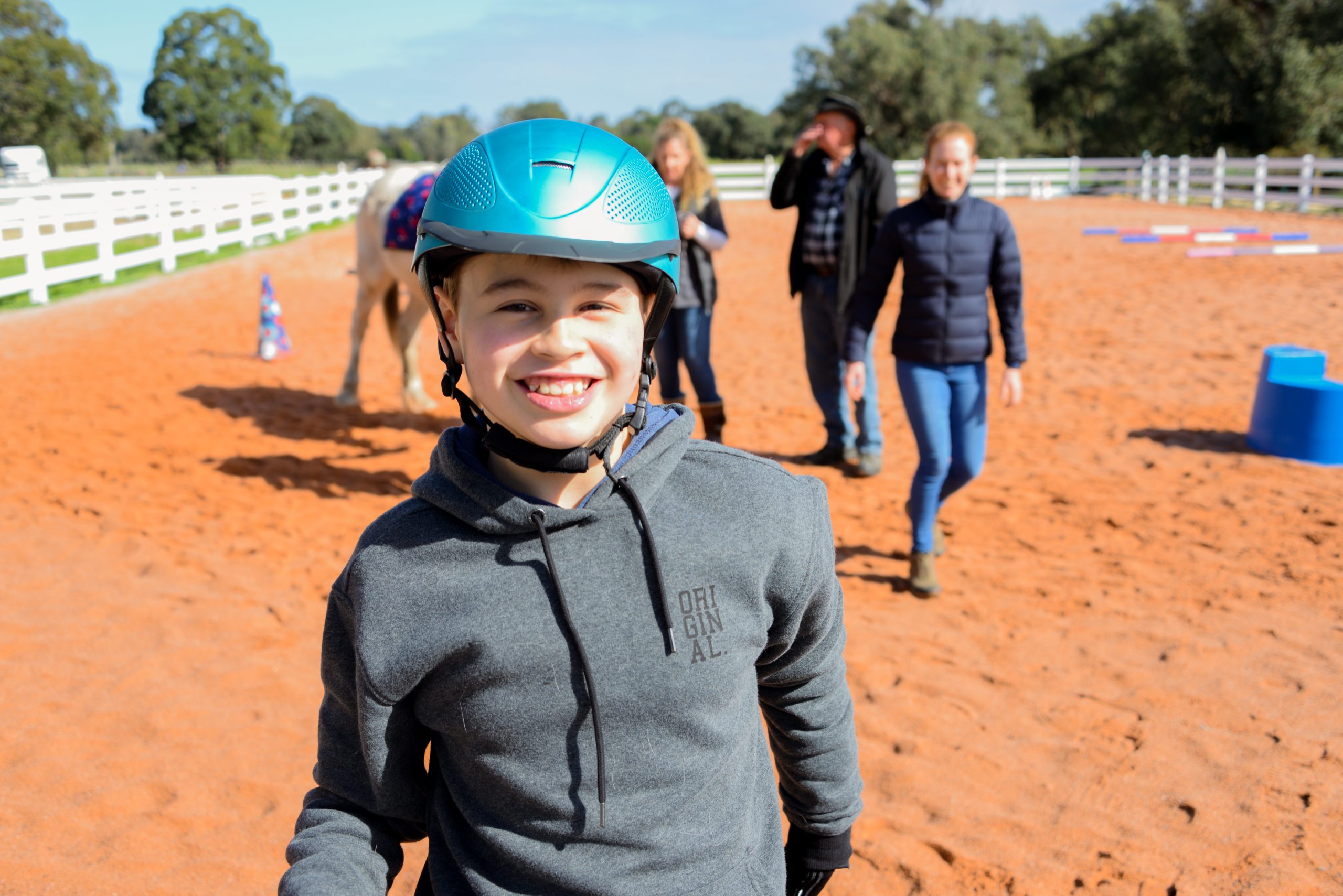 Young boy Anthony smiles at the camera wearing a blue horse riding helmet. In the background is an equistrian facility with a horse and several helpers walking beside it.