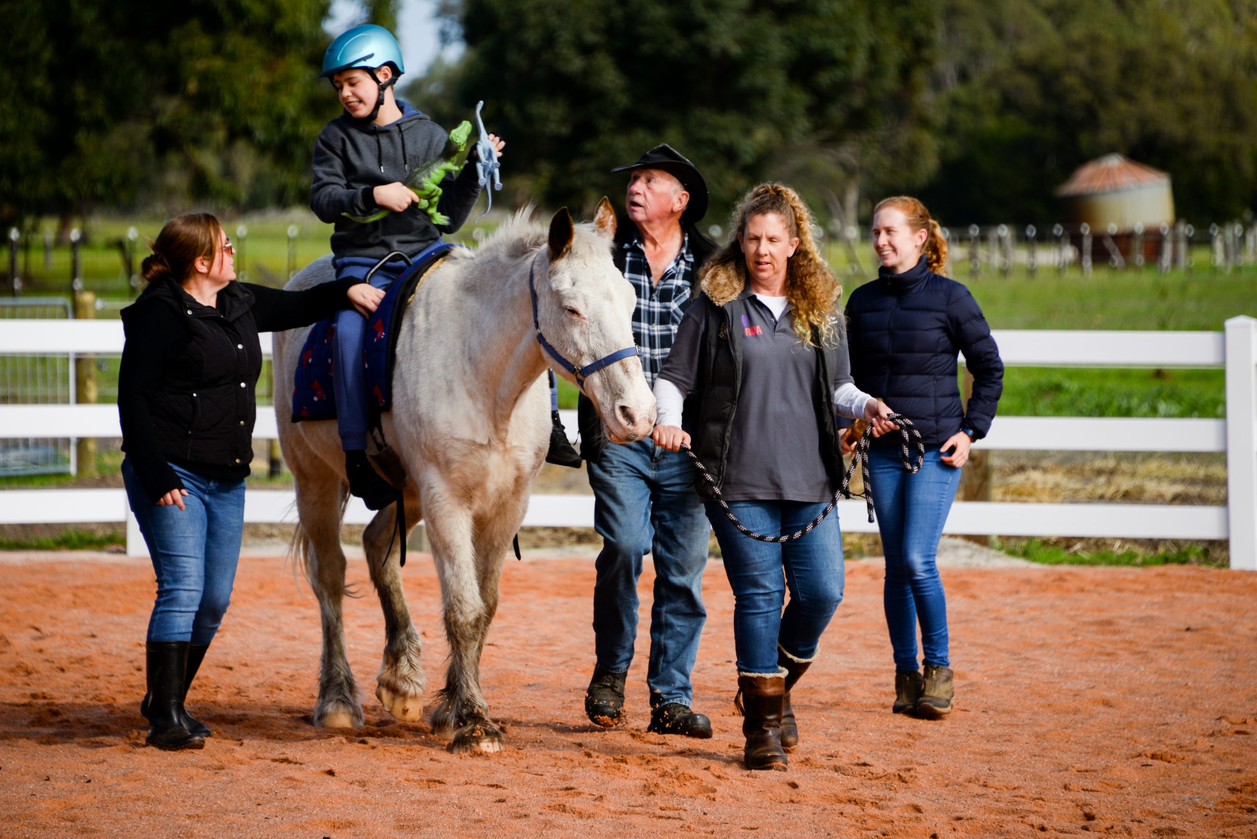 Young biy is sitting on a white horse during hippotherapy. He is surrounded by four people who are assisting