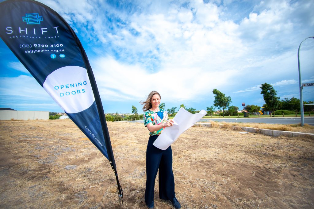 Image of a Rocky Bay staff member looking at building plans on a patch of land. There is a SHIFT Accessible Homes banner in the background.