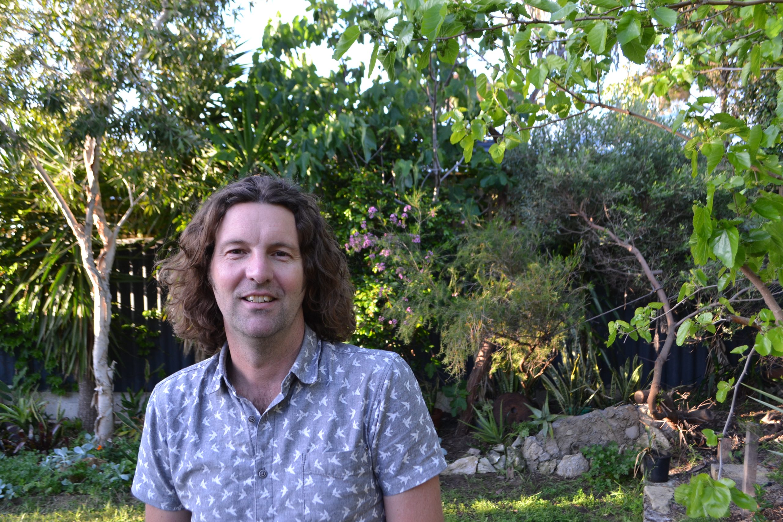 Glenn is smiling at the camera and is standing outside with lots of greenery in the background