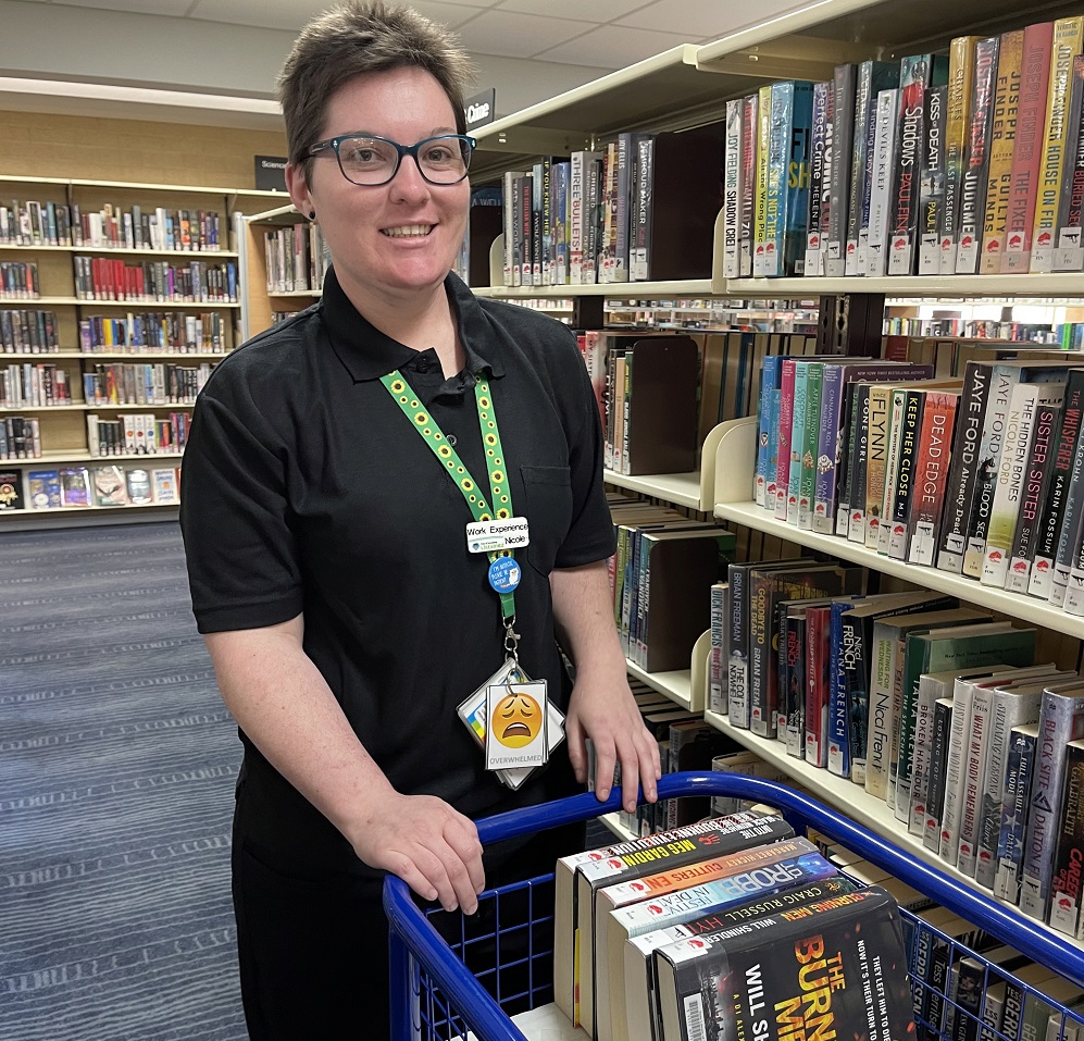 Nicole working at Woodvale Library. She is pushing a trolley of books and smiling at the camera.