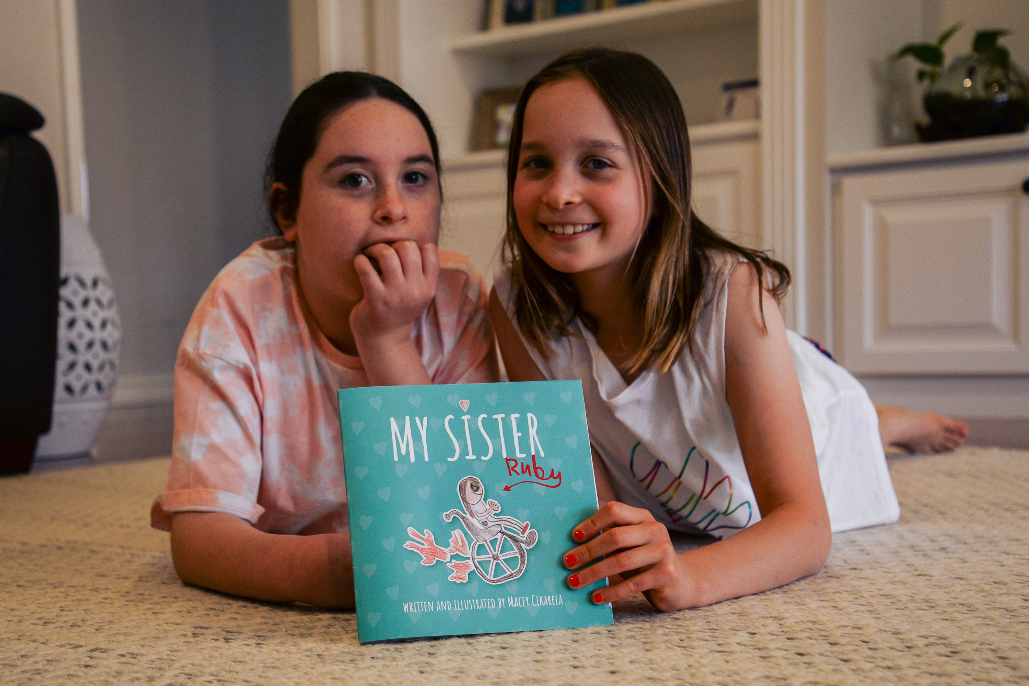 Sisters Ruby and Macey are laying on the floor holding Macey's children's book called 'My Sister'