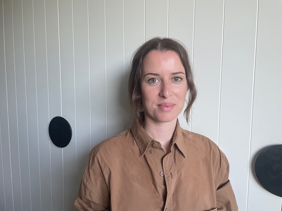 Gemma is standing in front of a white wall and smiling at the camera. She is wearing a brown shirt.