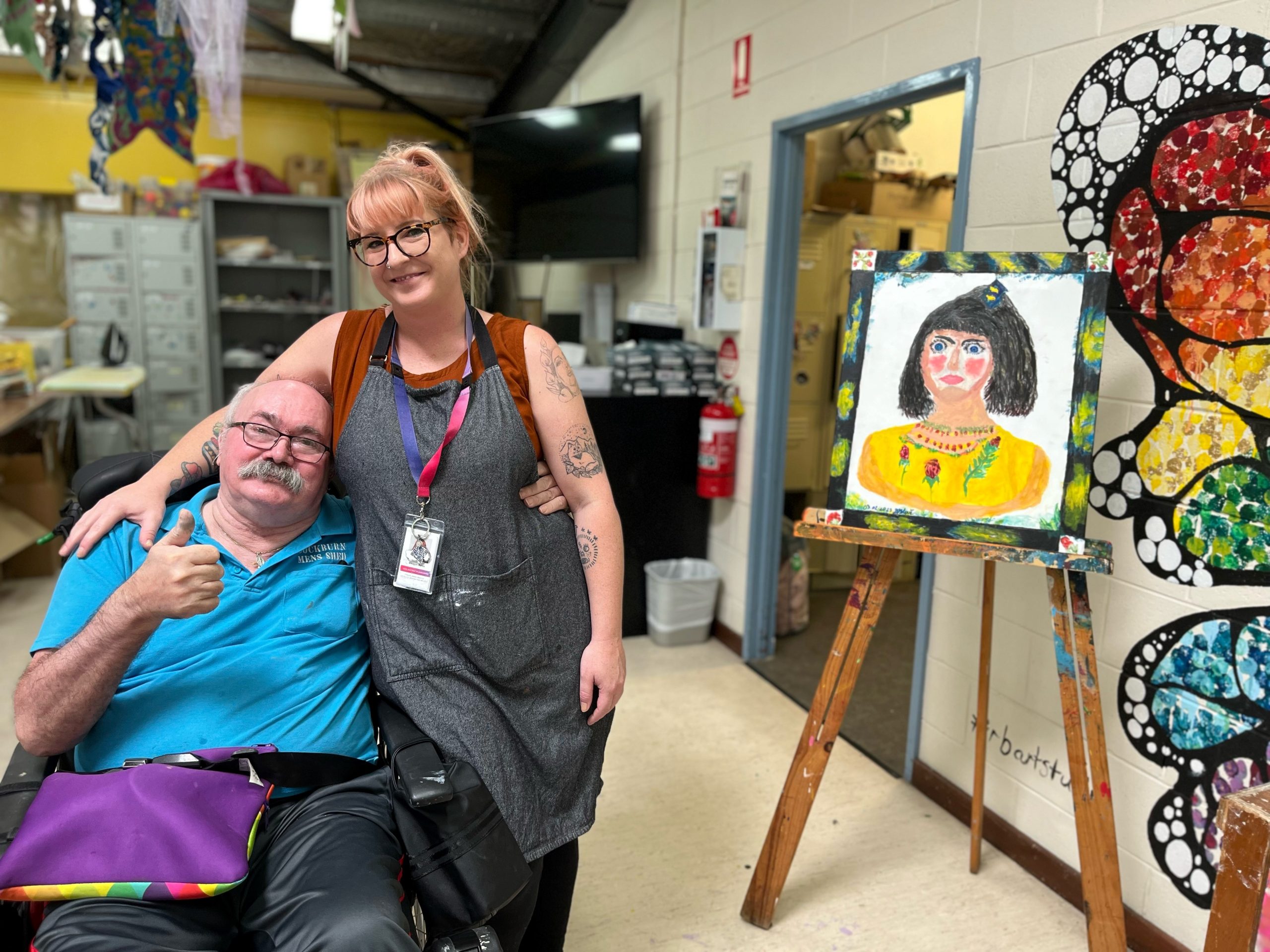 Artist Blaz with Rocky Bay Support Worker Meagan in the Studio. Blaz's colourful artwork if featured on an easel next to Meagan