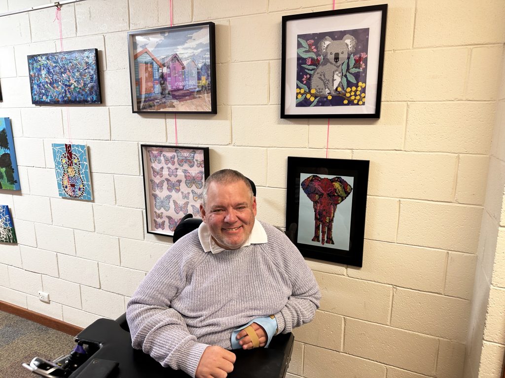 Brian is sitting infront of his diamond gem artwork. He wears a purple jumper and has a smile on his face.