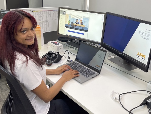 Julie Marie sits at her computer in the APM office. She is wearing a white shirt and smiling at the camera