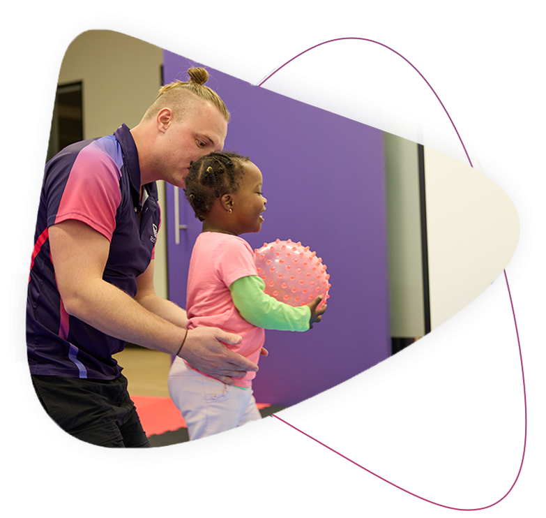 Rocky Bay Sprouts Kindy Readiness program. A young girl wearing a pink t-shirt is holding a pink ball and a Rocky Bay therapist is standing behind her supporting her