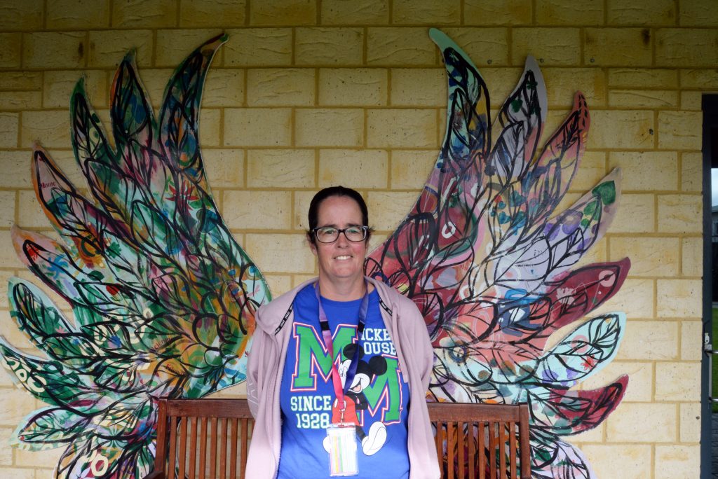 Support worker Sheree Pearce standing in front of the mural of colourful wings. She is wearing a purple Mickey Mouse shirt with a light pink jumper and her Rocky Bay lanyard on.