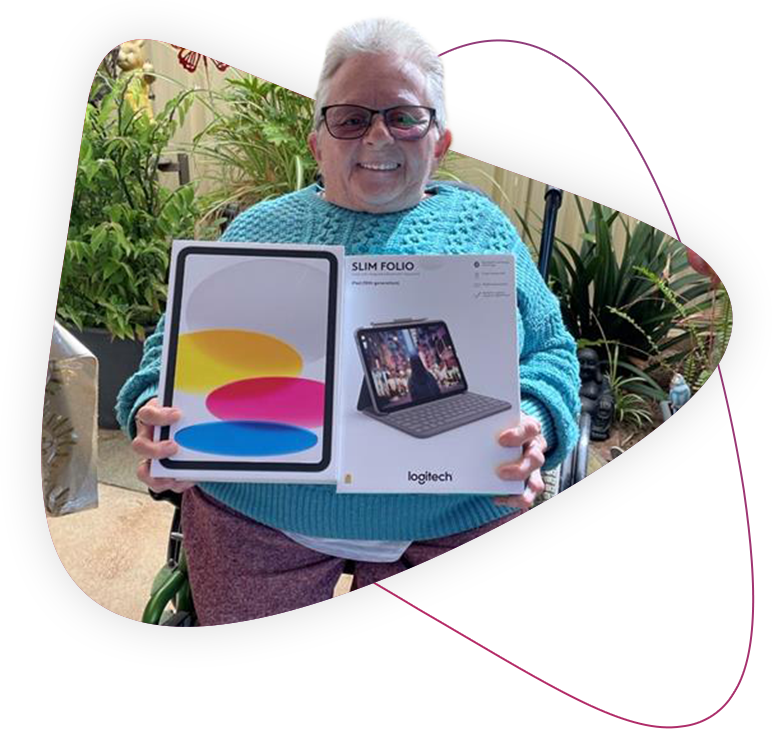 Customer Debbie, holding her new iPad and Keyboard accessory. She wears a blue knit jumper, wears glasses and has a smile on her face.