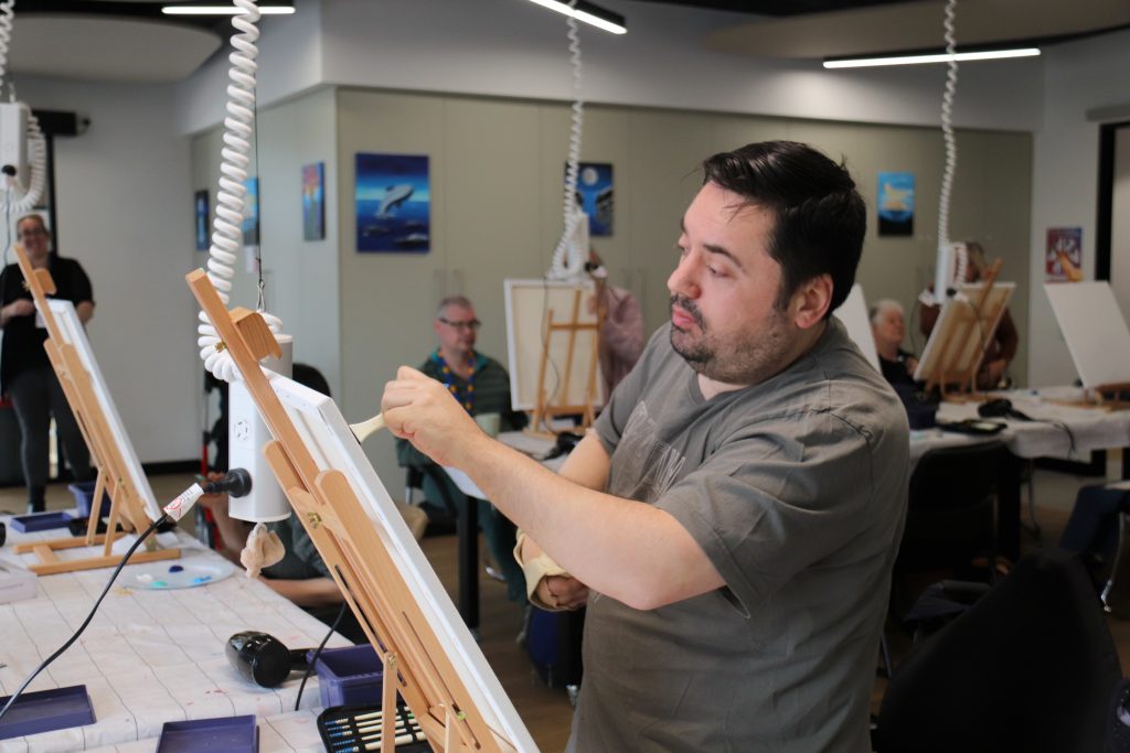 A man focused on painting on a canvas in a brightly lit room, with other artists in the background also working on their canvases.