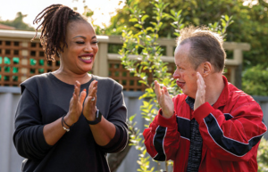 A photo of a Rocky Bay customer with his support worker. They are standing in the garden and looking at each other and smiling. They are clapping hands.