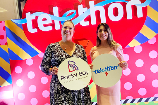 Rocky Bay staff members Kelly-Anne and Cristina at the Telethon grant breakfast. They are smiling at the camera and holding a sign with the Rocky Bay and Telethon logo.