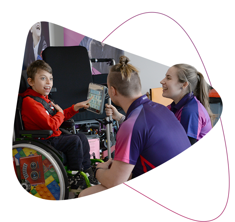 A photo of a young Rocky Bay customer sitting in his wheelchair and talking to two Rocky Bay therapists using his AAC device. He is looking at them and smiling while pointing at the screen of his device. His wheelchair has a colourful lego design on the wheel.