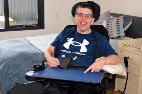 Rocky Bay customer Jason smiles for the camera. He is sitting in a wheelchair in his new Specialist Disability Accommodation room.