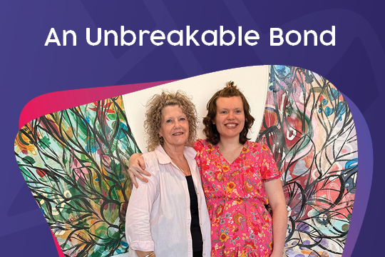 Lydia and her mum Leonie with their arms around each other in front of a pair of multi-colour angel wings wall art smiling at the camera